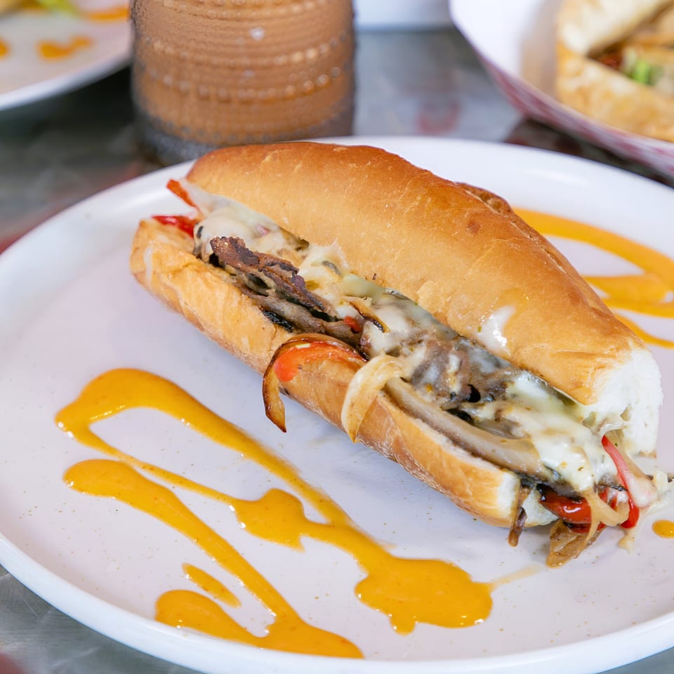 A Knockout Wings cheesesteak sandwich with melted cheese, steak, and peppers on a white plate with sauce.