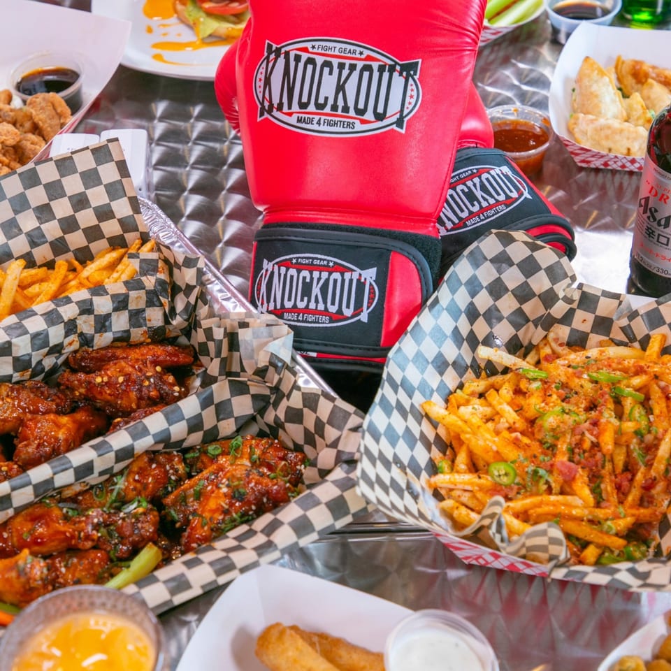 Knockout Wings' fast-food spread featuring wings, fries, and dumplings, with boxing gloves on a metal table.